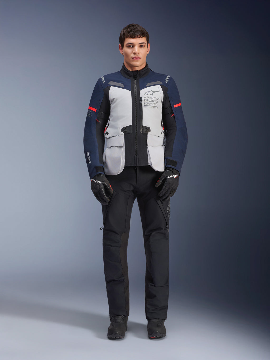 Kurtka Alpinestars ST-7 2L GORE-TEX, turystyczna kurtka motocyklowa adventure, kolor Ice Gray, granatowy i czarny, noszona z czarnymi spodniami i rękawicami, duże przednie kieszenie cargo, czerwone akcenty na rękawach, laminowana powłoka GORE-TEX zapewniająca wodoodporność.