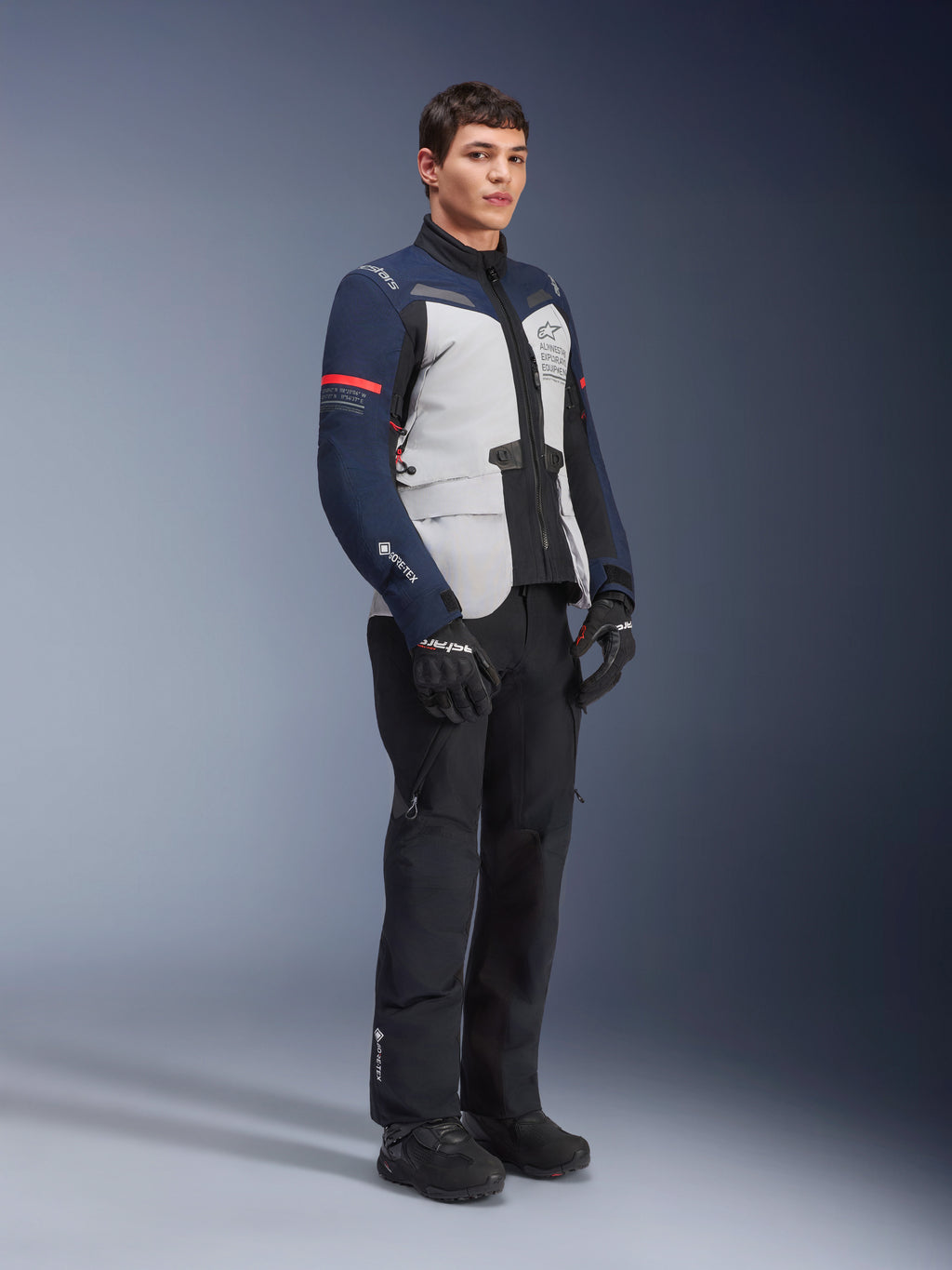 Kurtka Alpinestars ST-7 2L GORE-TEX w kolorystyce Ice Gray, Dark Blue i czarnej z czerwonymi akcentami, noszona przez modela z czarnymi spodniami motocyklowymi i rękawicami, profesjonalna odzież motocyklowa adventure touring