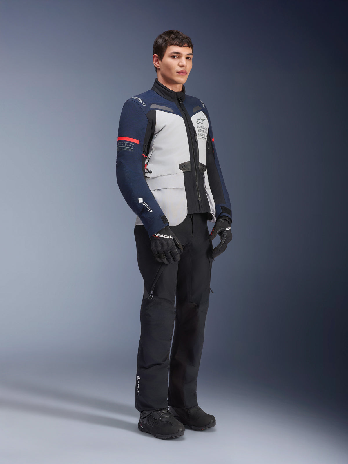 Kurtka Alpinestars ST-7 2L GORE-TEX w kolorystyce Ice Gray, Dark Blue i czarnej z czerwonymi akcentami, noszona przez modela z czarnymi spodniami motocyklowymi i rękawicami, profesjonalna odzież motocyklowa adventure touring