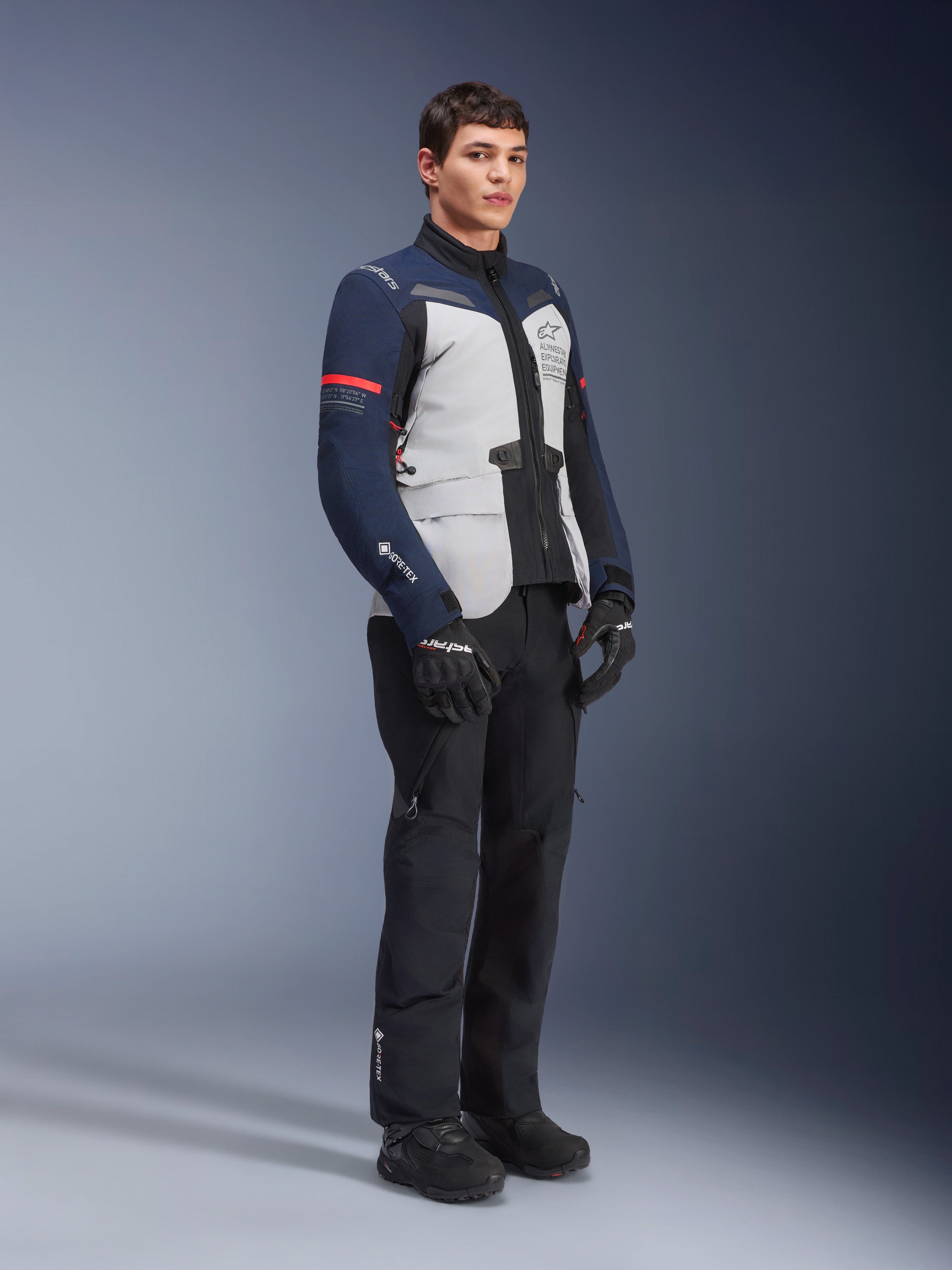 Kurtka Alpinestars ST-7 2L GORE-TEX w kolorystyce Ice Gray, Dark Blue i czarnej z czerwonymi akcentami, noszona przez modela z czarnymi spodniami motocyklowymi i rękawicami, profesjonalna odzież motocyklowa adventure touring