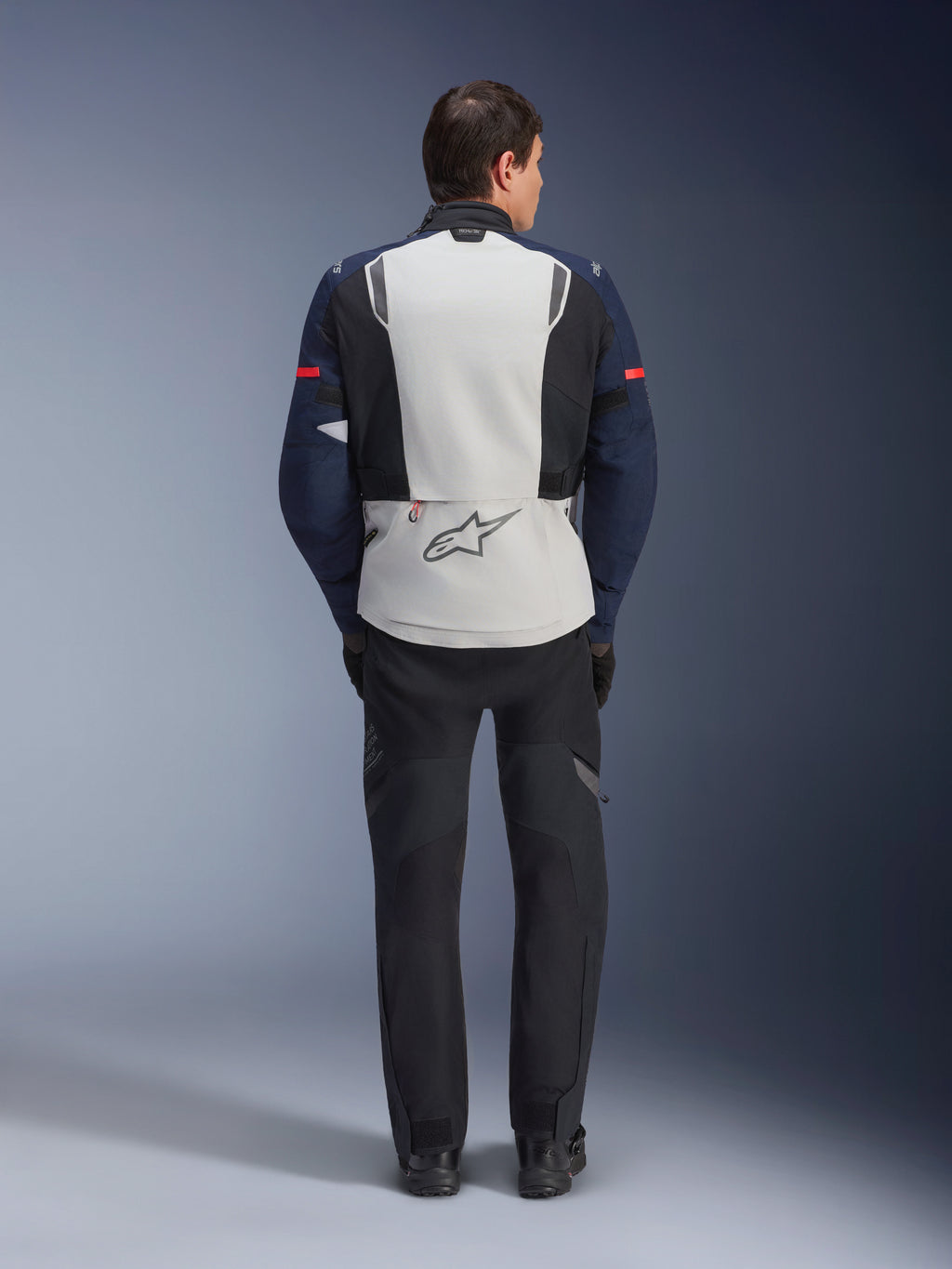 Kurtka Alpinestars ST-7 2L GORE-TEX, widok od tyłu motocyklisty w turystycznej kurtce motocyklowej typu adventure w kolorach Ice Gray, ciemnoniebieskim i czarnym, z dużym panelem na plecach z logo Alpinestars i dopasowanymi czarnymi spodniami, zaprojektowana do jazdy w każdych warunkach pogodowych.