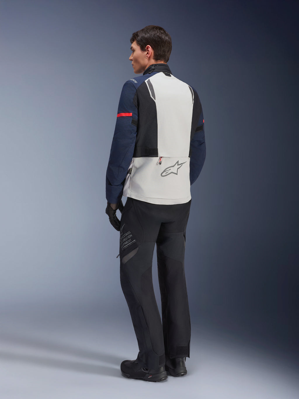 Model w kurtce Alpinestars ST-7 2L GORE-TEX w kolorze Ice Gray Dark Blue i czarnym, widok z tyłu, jasnoszary panel na plecach z logo Alpinestars, ciemnoniebieskie rękawy z czerwonym paskiem, czarne spodnie i buty adventure, motocyklowa odzież turystyczna
