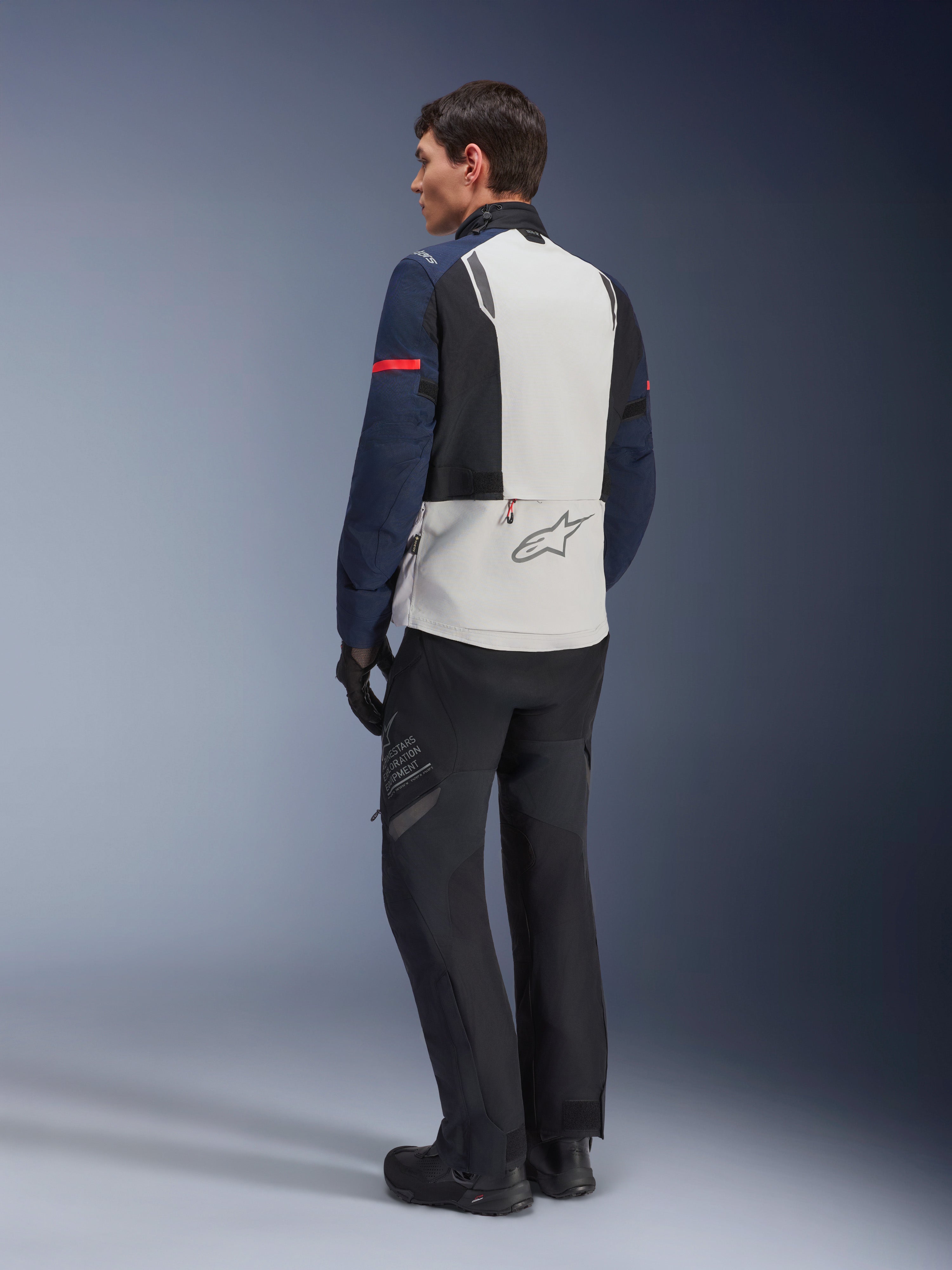 Model w kurtce Alpinestars ST-7 2L GORE-TEX w kolorze Ice Gray Dark Blue i czarnym, widok z tyłu, jasnoszary panel na plecach z logo Alpinestars, ciemnoniebieskie rękawy z czerwonym paskiem, czarne spodnie i buty adventure, motocyklowa odzież turystyczna