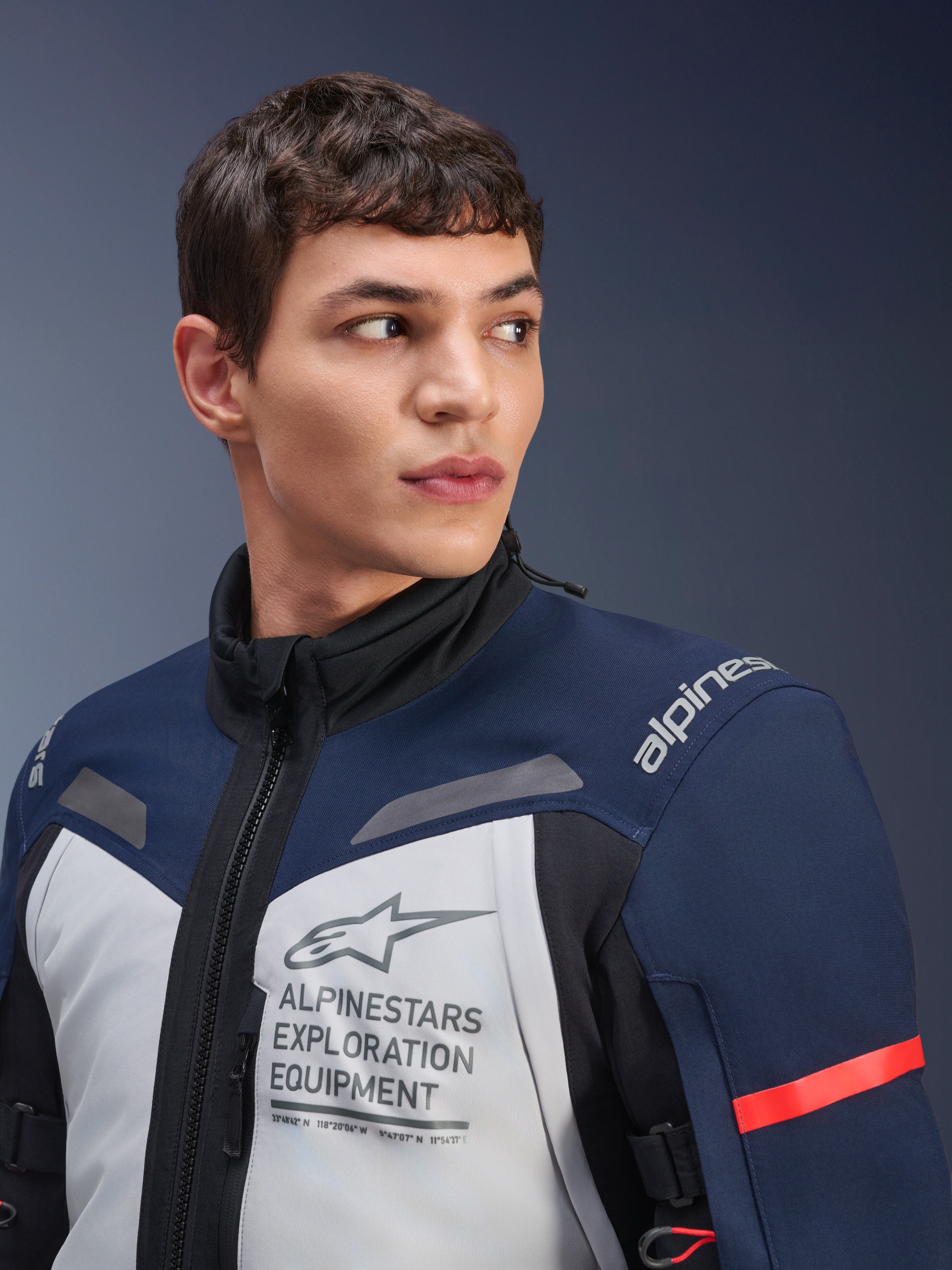 Kurtka Alpinestars ST-7 2L GORE-TEX, kolor Ice Gray, ciemnoniebieski i czarny, noszona przez modela, z logo Alpinestars Exploration Equipment na klatce piersiowej, wysokim kołnierzem i czerwonym akcentem na rękawie, wytrzymała konstrukcja tekstylna do turystyki adventure