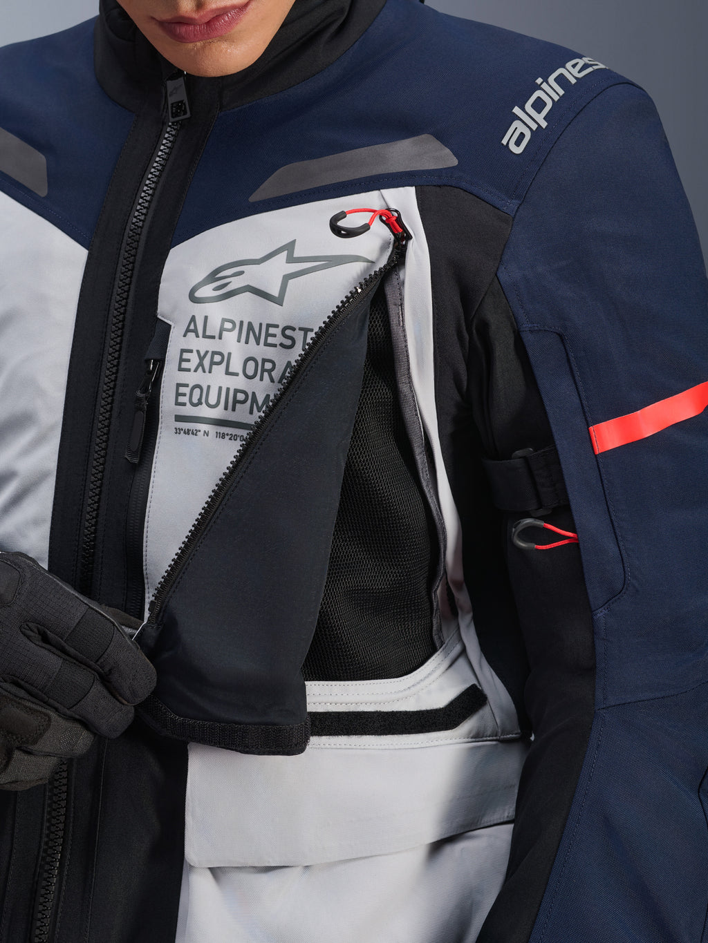 Kurtka Alpinestars ST-7 2L GORE-TEX, kurtka adventure touring, lodowy szary, granatowy i czarny, szczegółowy widok rozpiętego panelu wentylacyjnego na klatce piersiowej odsłaniającego czarną siatkę, logo Alpinestars Exploration Equipment i czerwone uchwyty zamków