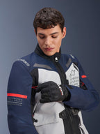 Kurtka Alpinestars ST-7 2L GORE-TEX, lodowoszara, ciemnoniebieska i czarna z pomarańczowymi akcentami, mężczyzna regulujący detal na rękawie, logo „Alpinestars Exploration Equipment” i współrzędne geograficzne, profesjonalna odzież motocyklowa adventure