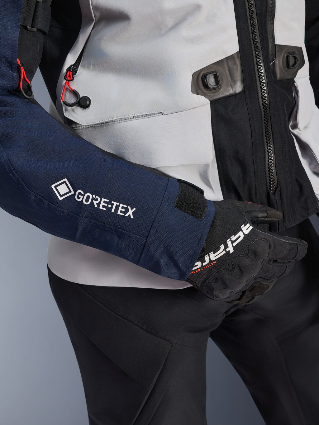 Kurtka Alpinestars ST-7 2L GORE-TEX, zbliżenie na ciemnoniebieski rękaw z białym logo GORE-TEX, korpus w kolorze lodowej szarości z czarnymi detalami i czerwonymi akcentami, czarne rękawice i spodnie Alpinestars, odzież motocyklowa adventure touring