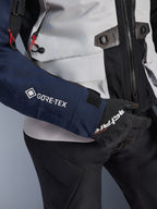 Kurtka Alpinestars ST-7 2L GORE-TEX, zbliżenie na ciemnoniebieski rękaw z białym logo GORE-TEX, korpus w kolorze lodowej szarości z czarnymi detalami i czerwonymi akcentami, czarne rękawice i spodnie Alpinestars, odzież motocyklowa adventure touring