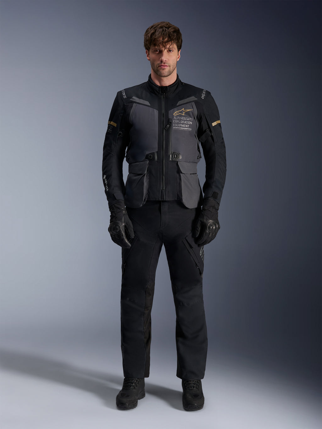 Męski model ubrany w kurtkę Alpinestars ST-7 2L GORE-TEX w kolorze Deep Gray Black Dark Khaki, z brandingiem „ALPINESTARS EXPLORATION EQUIPMENT” na klatce piersiowej, w połączeniu z pasującymi czarnymi spodniami motocyklowymi, rękawicami i butami, ujęcie studyjne od przodu.