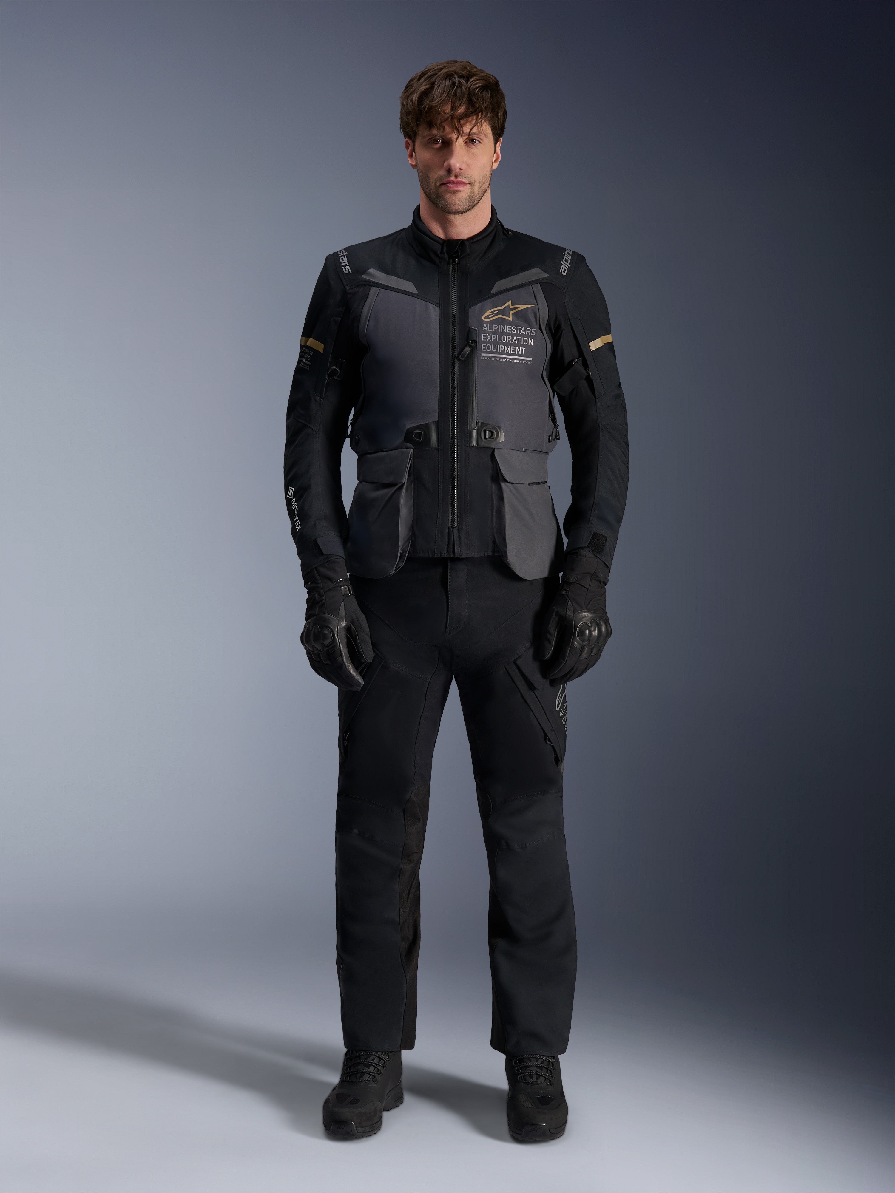 Męski model ubrany w kurtkę Alpinestars ST-7 2L GORE-TEX w kolorze Deep Gray Black Dark Khaki, z brandingiem „ALPINESTARS EXPLORATION EQUIPMENT” na klatce piersiowej, w połączeniu z pasującymi czarnymi spodniami motocyklowymi, rękawicami i butami, ujęcie studyjne od przodu.