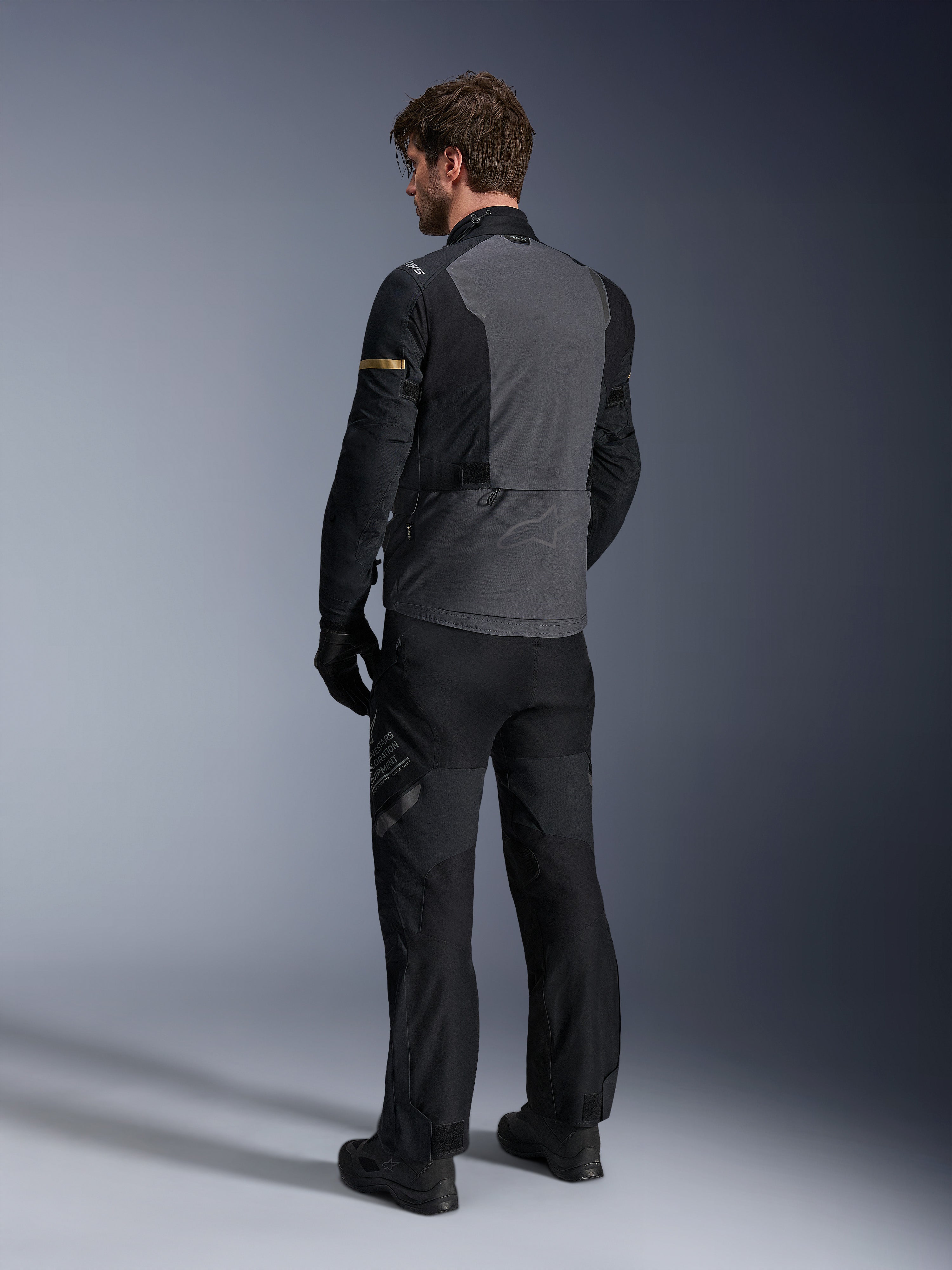 Kurtka Alpinestars ST-7 2L GORE-TEX, ciemnoszara i czarna z akcentami dark khaki, widok od tyłu motocyklisty w kurtce i pasujących spodniach motocyklowych, odzież adventure touring, wodoodporna laminowana konstrukcja