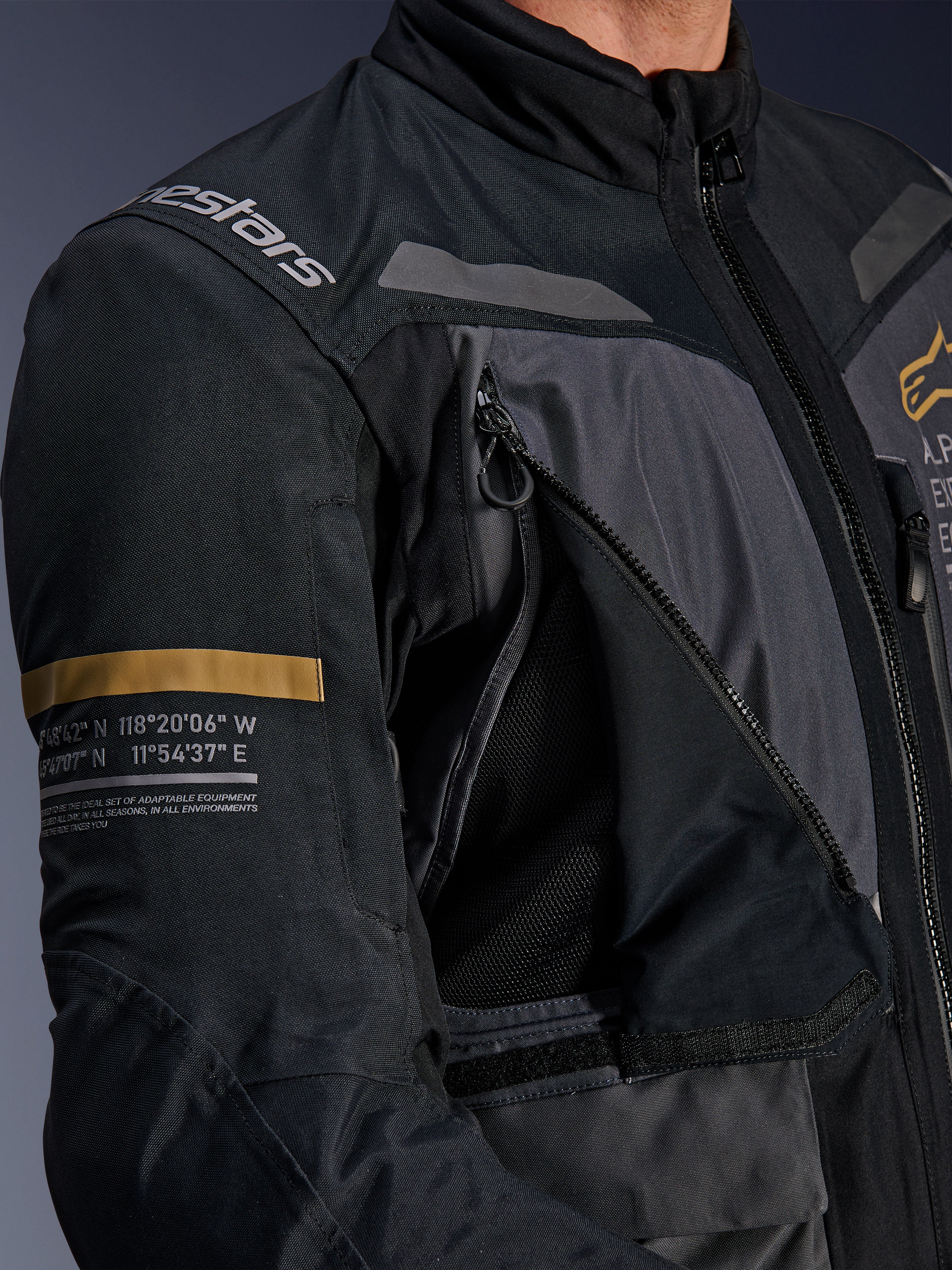 Kurtka Alpinestars ST-7 2L GORE-TEX, ciemnoszara i czarna, zbliżenie na otwarty panel wentylacyjny z siateczką na klatce piersiowej, logo Alpinestars na ramieniu, pasek w kolorze khaki na rękawie z nadrukowanymi współrzędnymi geograficznymi, techniczna odzież motocyklowa adventure-touring
