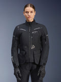 Kobieta ubrana w kurtkę Alpinestars Stella ST-7 2L GORE-TEX, czarna z ciemnoszarymi akcentami, motocyklowa kurtka turystyczna z dużymi kieszeniami cargo i wysokim kołnierzem, zestawiona z czarnymi rękawicami i spodniami, wodoodporna odzież do jazdy adventure
