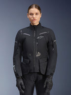 Kobieta ubrana w kurtkę Alpinestars Stella ST-7 2L GORE-TEX, czarna z ciemnoszarymi akcentami, motocyklowa kurtka turystyczna z dużymi kieszeniami cargo i wysokim kołnierzem, zestawiona z czarnymi rękawicami i spodniami, wodoodporna odzież do jazdy adventure