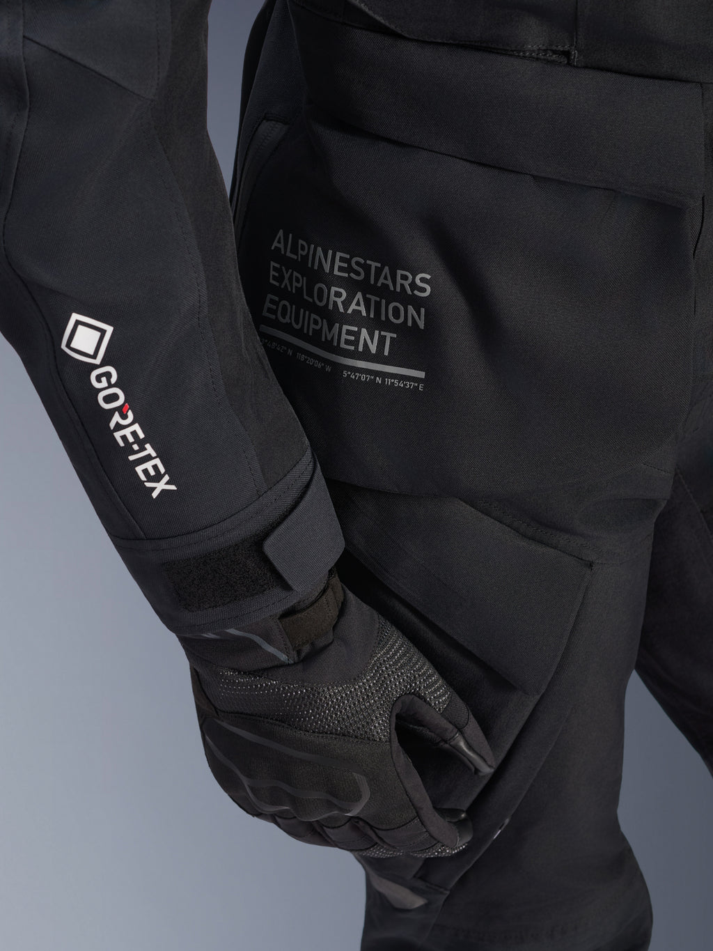 Damska kurtka Alpinestars Stella ST-7 2L GORE-TEX, czarna kurtka motocyklowa typu adventure touring, widok z bliska na rękaw z białym logo GORE-TEX, napis Alpinestars Exploration Equipment na biodrze, czarna rękawica motocyklowa, wodoodporny materiał tekstylny z funkcjonalnymi kieszeniami cargo