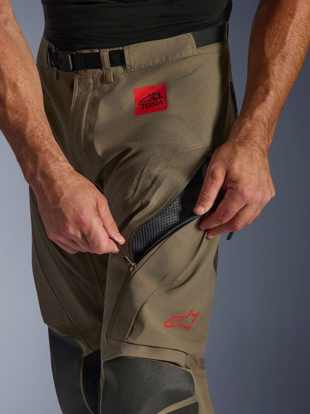 Alpinestars Nazca 3L GORE-TEX PRO Pants w kolorze Walnut Black, zbliżenie na detal uda z dłonią otwierającą duży wlot wentylacyjny na zamek, czerwona naszywka ALL TERRA i logo Alpinestars, techniczna odzież motocyklowa adventure