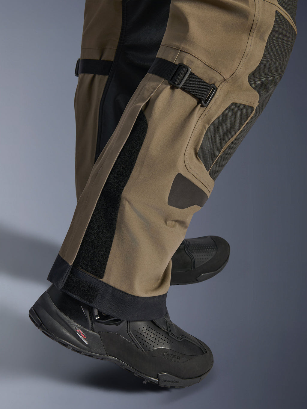 Zbliżenie na spodnie Alpinestars Nazca 3L GORE-TEX PRO w kolorze Walnut Black, ukazujące techniczne detale dolnej części nogawki, w tym regulatory objętości łydki i wzmocnienia Superfabric, noszone z czarnymi butami motocyklowymi adventure.