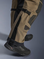 Zbliżenie na spodnie Alpinestars Nazca 3L GORE-TEX PRO w kolorze Walnut Black, ukazujące techniczne detale dolnej części nogawki, w tym regulatory objętości łydki i wzmocnienia Superfabric, noszone z czarnymi butami motocyklowymi adventure.