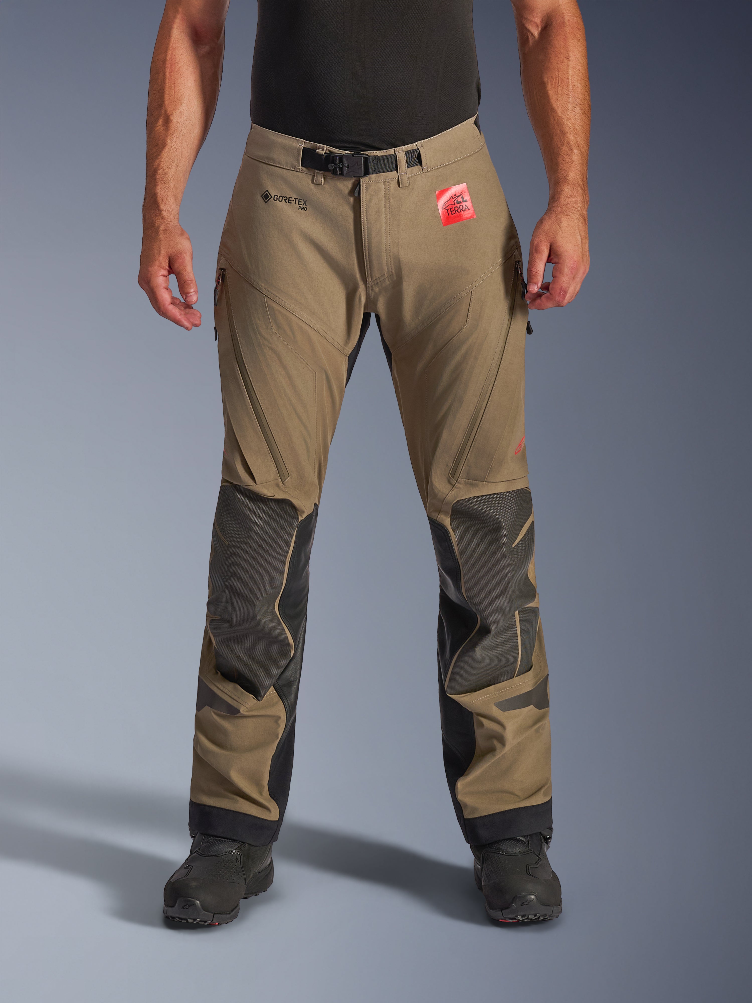 Alpinestars Nazca 3L GORE-TEX PRO Pants w kolorze Walnut Black, męskie spodnie turystyczne Adventure, brązowy materiał z czarnymi wzmocnieniami Superfabric na kolanach, zapinane na zamek wywietrzniki na udach, magnetyczne zapięcie w pasie, widok z przodu na modelu w czarnych butach, trwała wodoodporna odzież motocyklowa