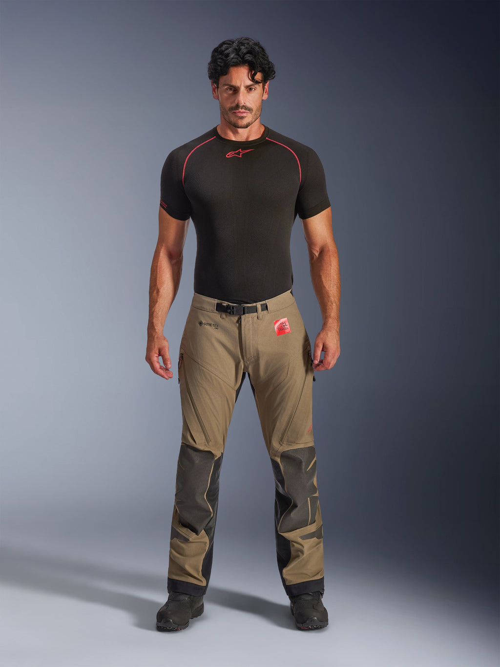 Model w spodniach Alpinestars Nazca 3L GORE-TEX PRO w kolorze orzechowego brązu i czerni, techniczna odzież motocyklowa typu adventure ze wzmocnieniami kolan Superfabric i magnetyczną klamrą w pasie, w połączeniu z czarną koszulką termoaktywną Alpinestars, widok z przodu w studio.