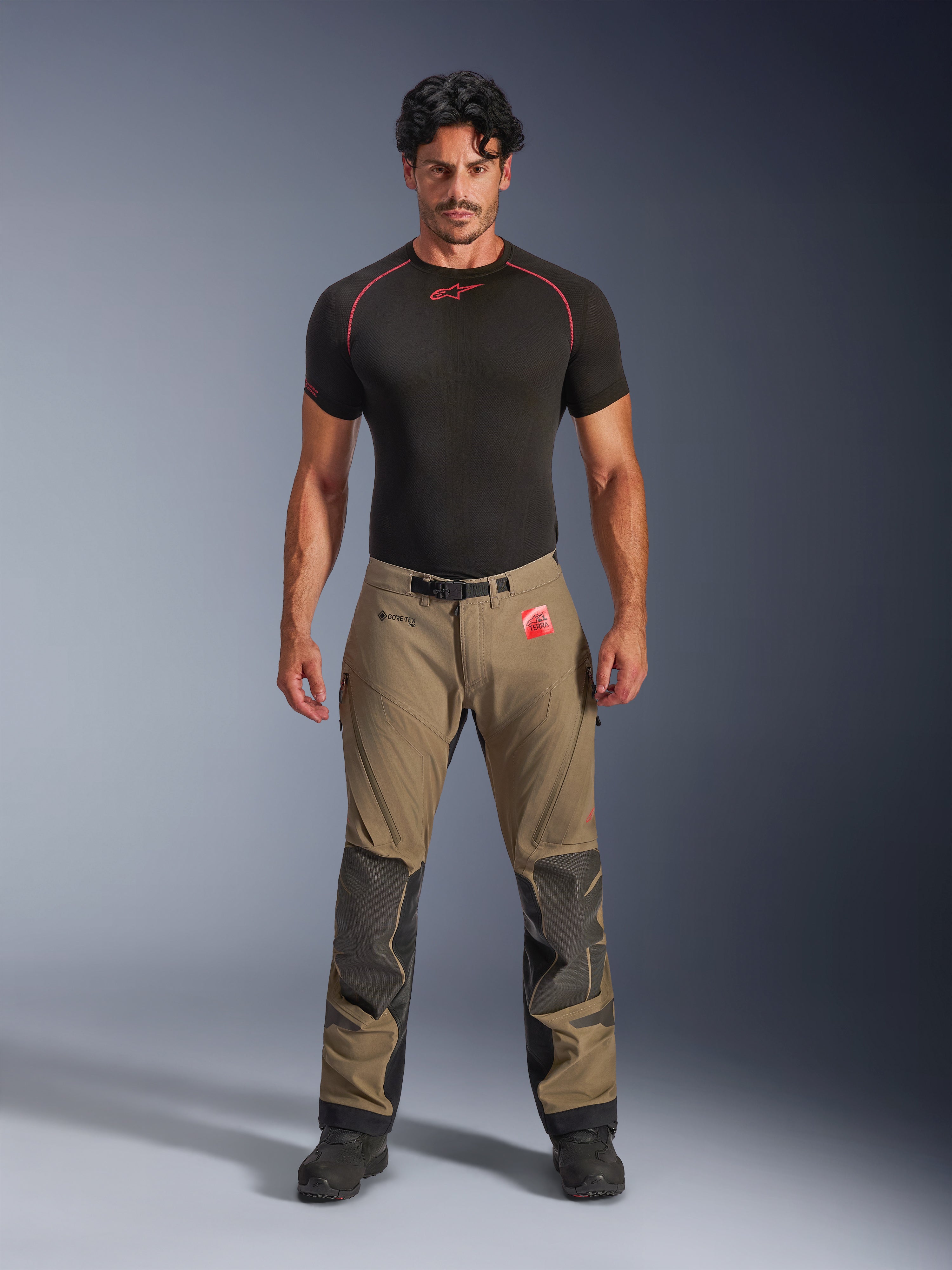 Model w spodniach Alpinestars Nazca 3L GORE-TEX PRO w kolorze orzechowego brązu i czerni, techniczna odzież motocyklowa typu adventure ze wzmocnieniami kolan Superfabric i magnetyczną klamrą w pasie, w połączeniu z czarną koszulką termoaktywną Alpinestars, widok z przodu w studio.