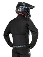 Widok od tyłu motocyklisty ubranego w czarną kurtkę Alpinestars Pro-Dura, wytrzymała tekstylna kurtka enduro z ripstopu, z dużą tylną kieszenią cargo i szarym napisem Alpinestars w dolnej części pleców, w zestawie z czarnym kaskiem motocrossowym.