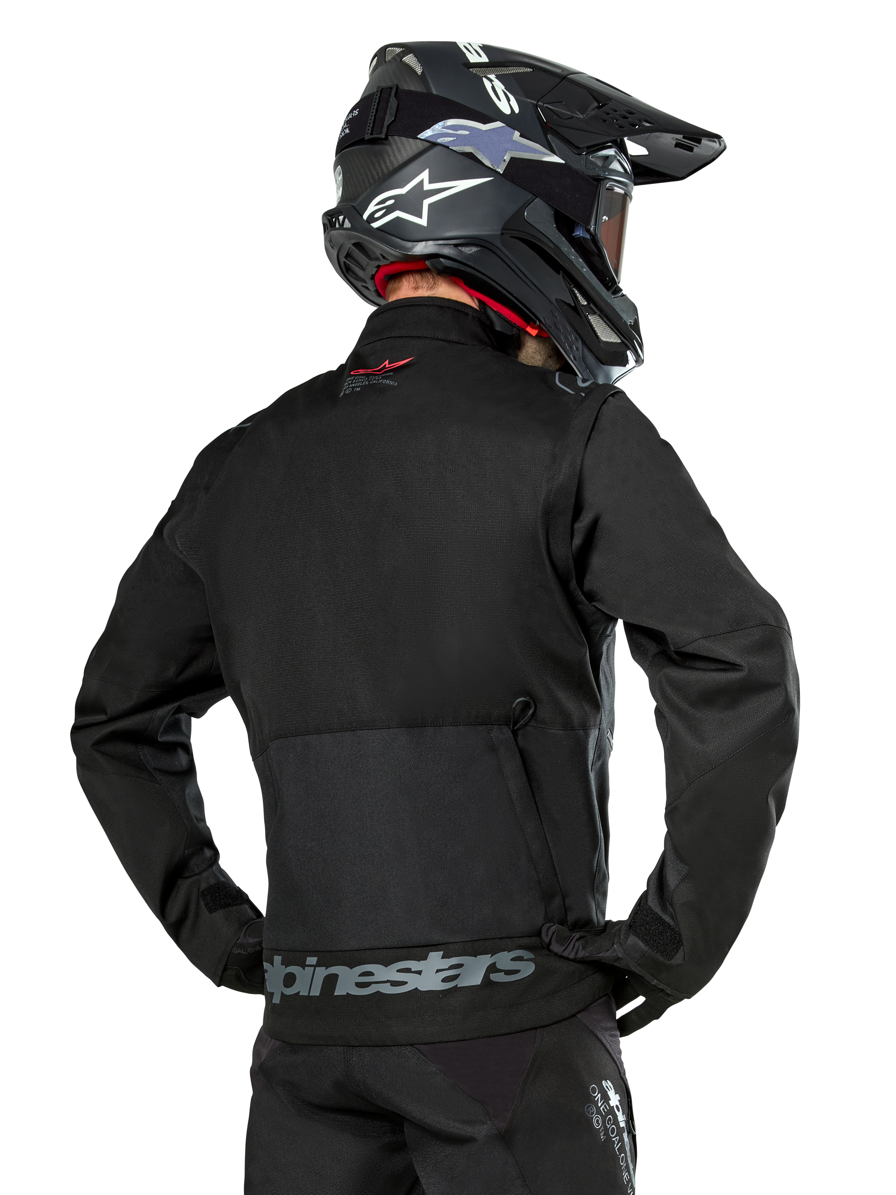 Widok od tyłu motocyklisty ubranego w czarną kurtkę Alpinestars Pro-Dura, wytrzymała tekstylna kurtka enduro z ripstopu, z dużą tylną kieszenią cargo i szarym napisem Alpinestars w dolnej części pleców, w zestawie z czarnym kaskiem motocrossowym.