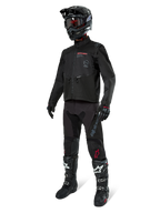 Motocyklista motocrossowy w pełnym stroju wyścigowym, kurtka Alpinestars Pro-Dura w kolorze czarnym, czarne spodnie z szarym brandingiem i czerwonymi akcentami, czarny kask z goglami oraz czarno-białe buty motocrossowe, dopasowany ubiór do jazdy enduro i off-road