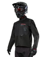 Alpinestars Pro-Dura Jacket, motocyklowa kurtka off-road, czarna z czerwonymi i szarymi elementami graficznymi, posiada przednie kieszenie na zamek i wysoki kołnierz, zestawiona z czarnym kaskiem motocrossowym i rękawicami do jazdy enduro