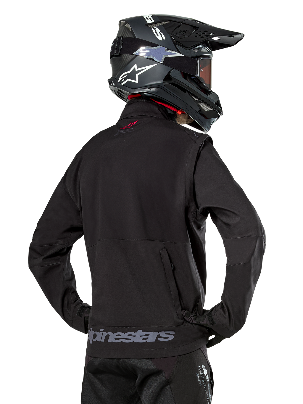 Kurtka Alpinestars Lite-Dura Softshell, czarna, widok z tyłu na motocykliście, szary napis Alpinestars na dolnym brzegu i małe czerwone logo na kołnierzu, zestaw z czarnym kaskiem motocrossowym, do jazdy enduro i off-road