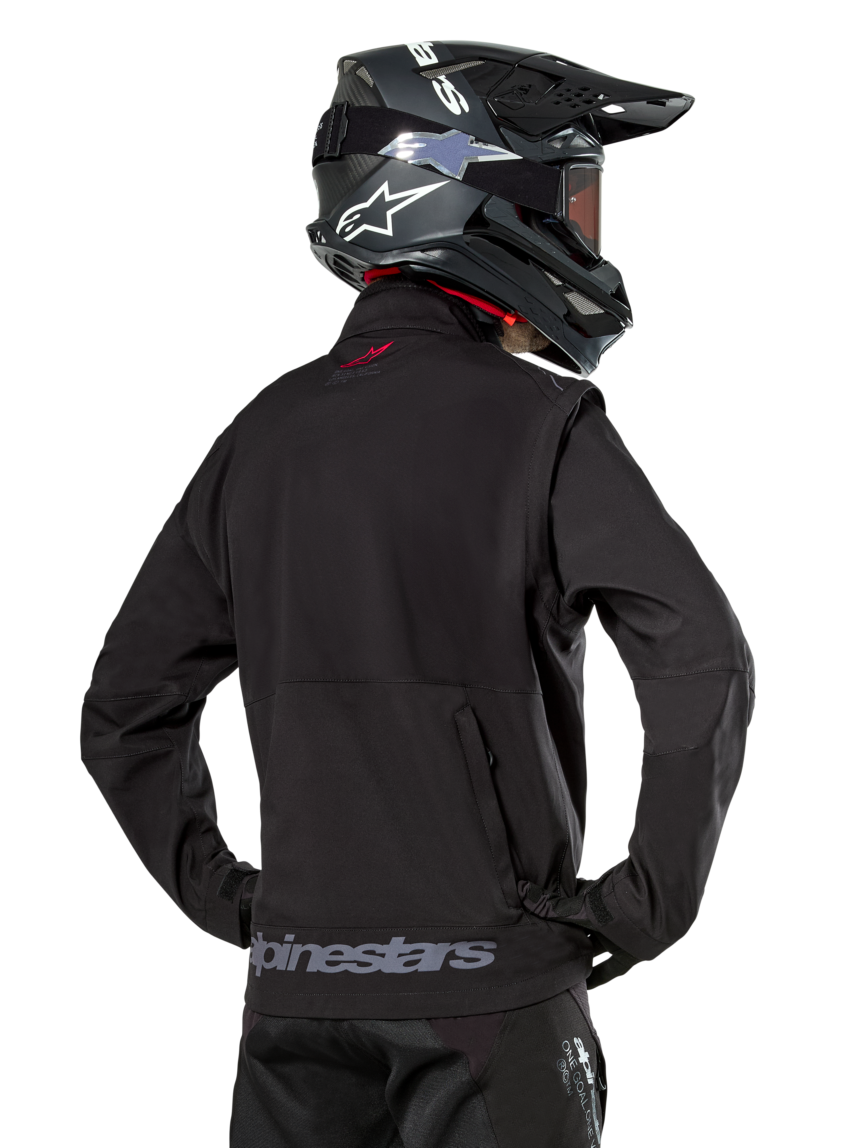 Kurtka Alpinestars Lite-Dura Softshell, czarna, widok z tyłu na motocykliście, szary napis Alpinestars na dolnym brzegu i małe czerwone logo na kołnierzu, zestaw z czarnym kaskiem motocrossowym, do jazdy enduro i off-road