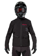 Motocyklista motocrossowy w kurtce Alpinestars Lite-Dura Softshell w kolorze czarnym, czerwone i szare detale logo na piersi, czarny kask z czerwonymi goglami, czarne rękawice i spodnie, widok z przodu technicznej odzieży off-road