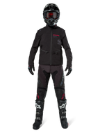 Motocyklista motocrossowy w kurtce Alpinestars Lite-Dura Softshell w kolorze czarnym z czerwonymi detalami logo, dopasowane czarne spodnie off-road, czarny kask oraz biało-czarne buty, kompletny profesjonalny zestaw wyścigowy enduro