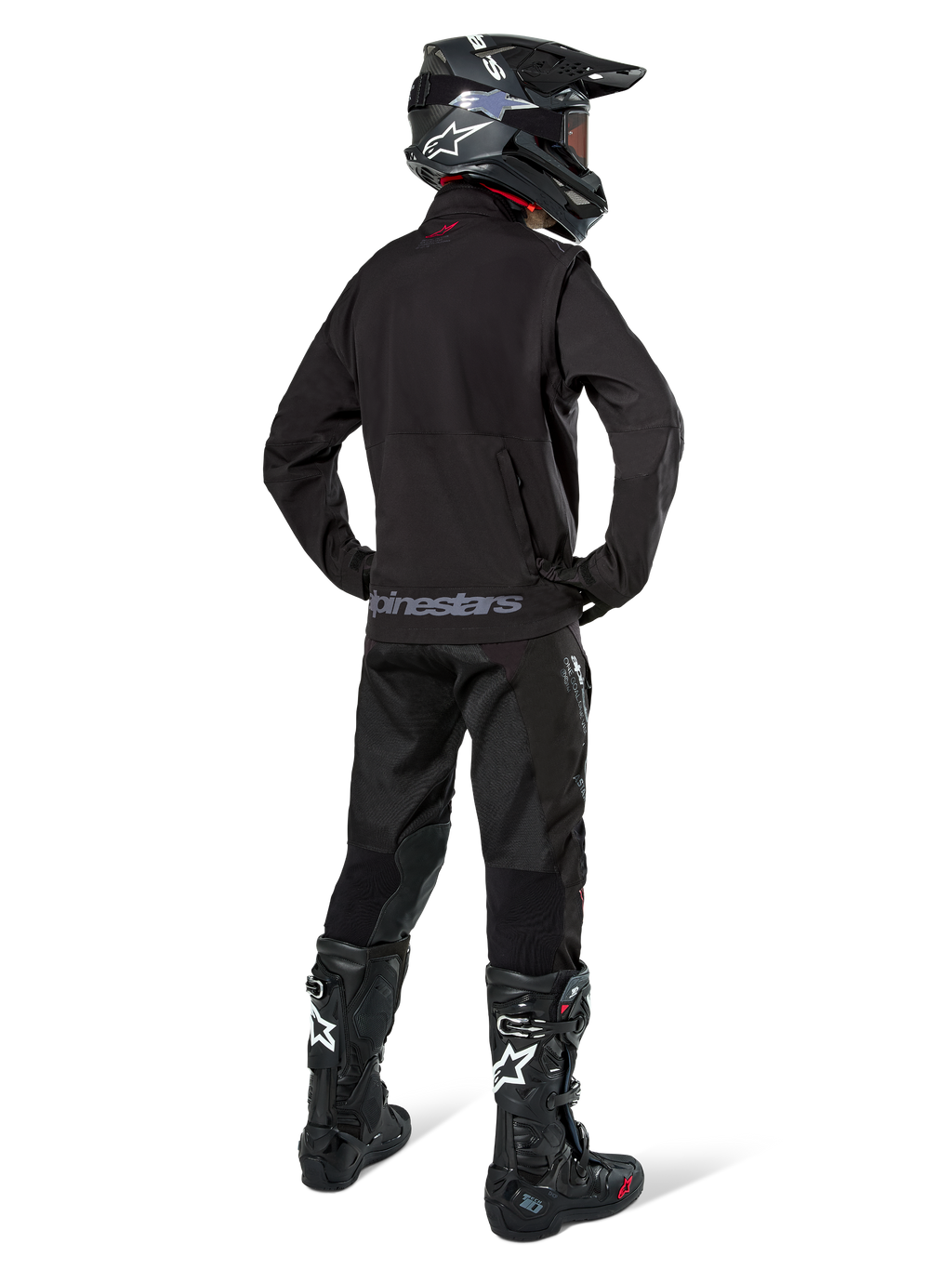 Zawodnik enduro widziany od tyłu w czarnej kurtce Alpinestars Lite-Dura Softshell, zestawiony z czarnymi technicznymi spodniami i butami motocyklowymi, z tylną kieszenią na zamek i logo Alpinestars na dolnym brzegu, kompletny strój off-road do motocrossu.