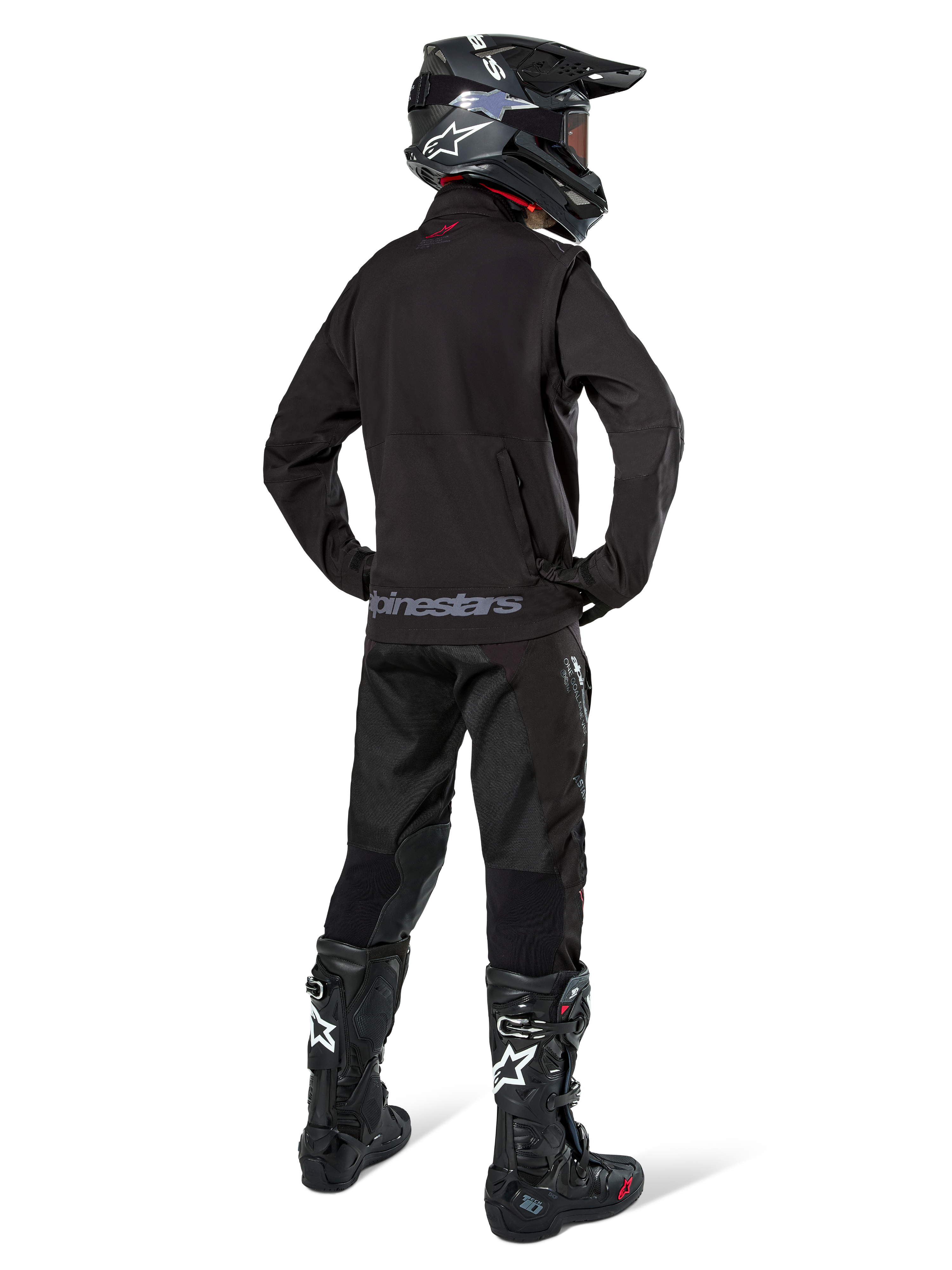 Zawodnik enduro widziany od tyłu w czarnej kurtce Alpinestars Lite-Dura Softshell, zestawiony z czarnymi technicznymi spodniami i butami motocyklowymi, z tylną kieszenią na zamek i logo Alpinestars na dolnym brzegu, kompletny strój off-road do motocrossu.