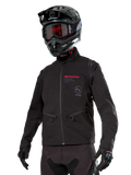Kurtka Alpinestars Lite-Dura Softshell, czarna terenowa kurtka motocyklowa z czerwonym brandingiem i logo AS, noszona z czarnym kaskiem motocrossowym, czerwonymi goglami i czarnymi rękawicami, kieszenie na ręce zapinane na zamek, regulowane mankiety, przeznaczona do jazdy enduro