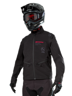 Kurtka Alpinestars Lite-Dura Softshell, czarna terenowa kurtka motocyklowa z czerwonym brandingiem i logo AS, noszona z czarnym kaskiem motocrossowym, czerwonymi goglami i czarnymi rękawicami, kieszenie na ręce zapinane na zamek, regulowane mankiety, przeznaczona do jazdy enduro