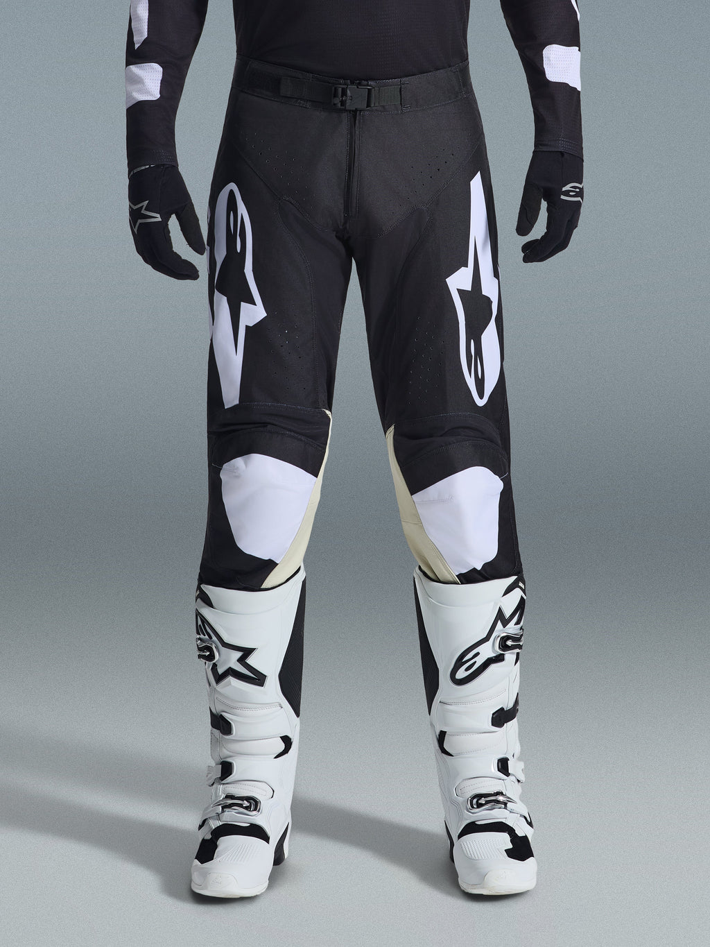 Alpinestars 2026 Racer Air Riway Pants, spodnie motocrossowe, czarno-białe, z wyraźnymi białymi logotypami Alpinestars na udach i panelach kolanowych, w zestawieniu z biało-czarnymi butami motocrossowymi i czarnymi rękawicami, sportowy krój race-fit do jazdy off-road