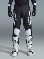 Alpinestars 2026 Racer Air Riway Pants, spodnie motocrossowe, czarno-białe, z wyraźnymi białymi logotypami Alpinestars na udach i panelach kolanowych, w zestawieniu z biało-czarnymi butami motocrossowymi i czarnymi rękawicami, sportowy krój race-fit do jazdy off-road