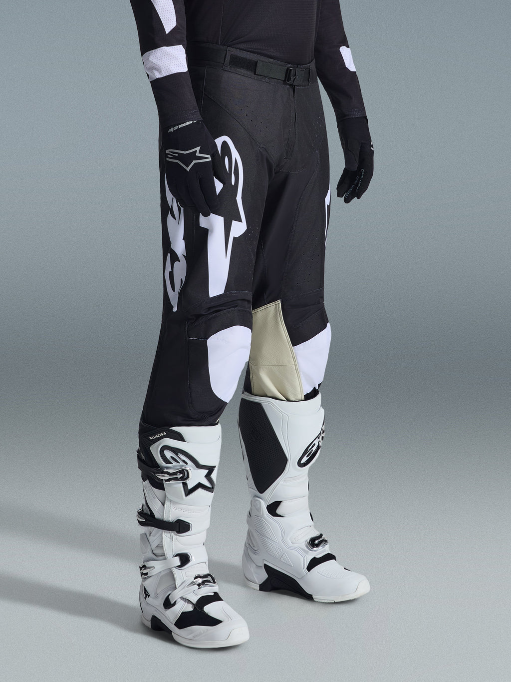 Alpinestars Racer Air Riway Pants 2026, spodnie motocrossowe, czarno-białe, zestawione z biało-czarnymi butami Tech Series i czarną bluzą z długim rękawem, białe logo Alpinestars, laserowo wycinane perforacje oraz jasnobrązowe panele z koziej skóry po wewnętrznej stronie kolan.