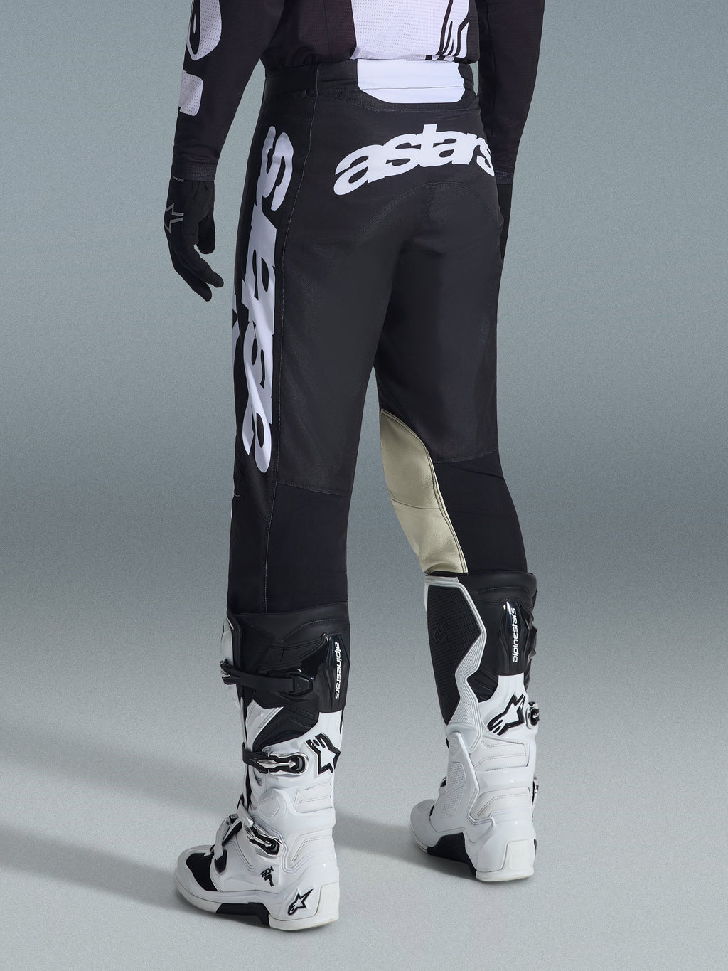 Alpinestars Racer Air Riway Pants 2026, spodnie motocrossowe, czarno-białe, widok z tyłu z dużym białym logo Astars na siedzeniu i nogawkach, beżowe skórzane panele po wewnętrznej stronie kolan, zestawione z biało-czarnymi butami Tech 7, wysokiej jakości odzież wyścigowa off-road.