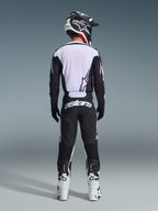 Motocyklista motocrossowy widziany od tyłu w spodniach Alpinestars 2026 Racer Air Riway w kolorze czarno-białym, dopasowanej czarno-białej bluzie z długim rękawem, biało-czarnych butach wyścigowych i czarnym kasku z logo Alpinestars