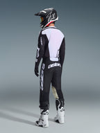 Motocyklista motocrossowy widziany od tyłu, ubrany w spodnie Alpinestars 2026 Racer Air Riway w kolorze czarno-białym, w zestawie z pasującą wentylowaną koszulką, czarno-białym kaskiem i białymi butami motocrossowymi, z widocznym białym brandingiem „astars” na siedzeniu.