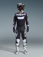 Zawodnik motocrossowy w spodniach Alpinestars 2026 Racer Air Riway w kolorze czarno-białym, pasująca czarno-biała koszulka z logo „astars”, czarny kask, gogle z iryzującymi szybkami i białe buty wyścigowe, kompletny profesjonalny zestaw odzieży off-road