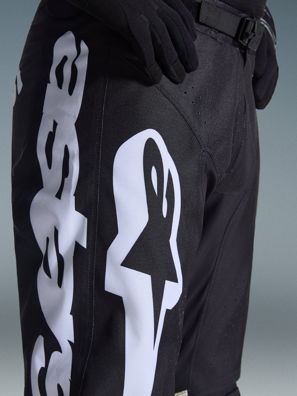 Alpinestars 2026 Racer Air Riway Pants, czarne spodnie motocrossowe z wyraźnym białym logo Alpinestars na udzie, zbliżenie pokazujące techniczny materiał i regulowany pasek w talii, noszone z czarnymi rękawicami do jazdy terenowej