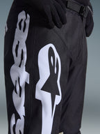 Alpinestars 2026 Racer Air Riway Pants, czarne spodnie motocrossowe z wyraźnym białym logo Alpinestars na udzie, zbliżenie pokazujące techniczny materiał i regulowany pasek w talii, noszone z czarnymi rękawicami do jazdy terenowej