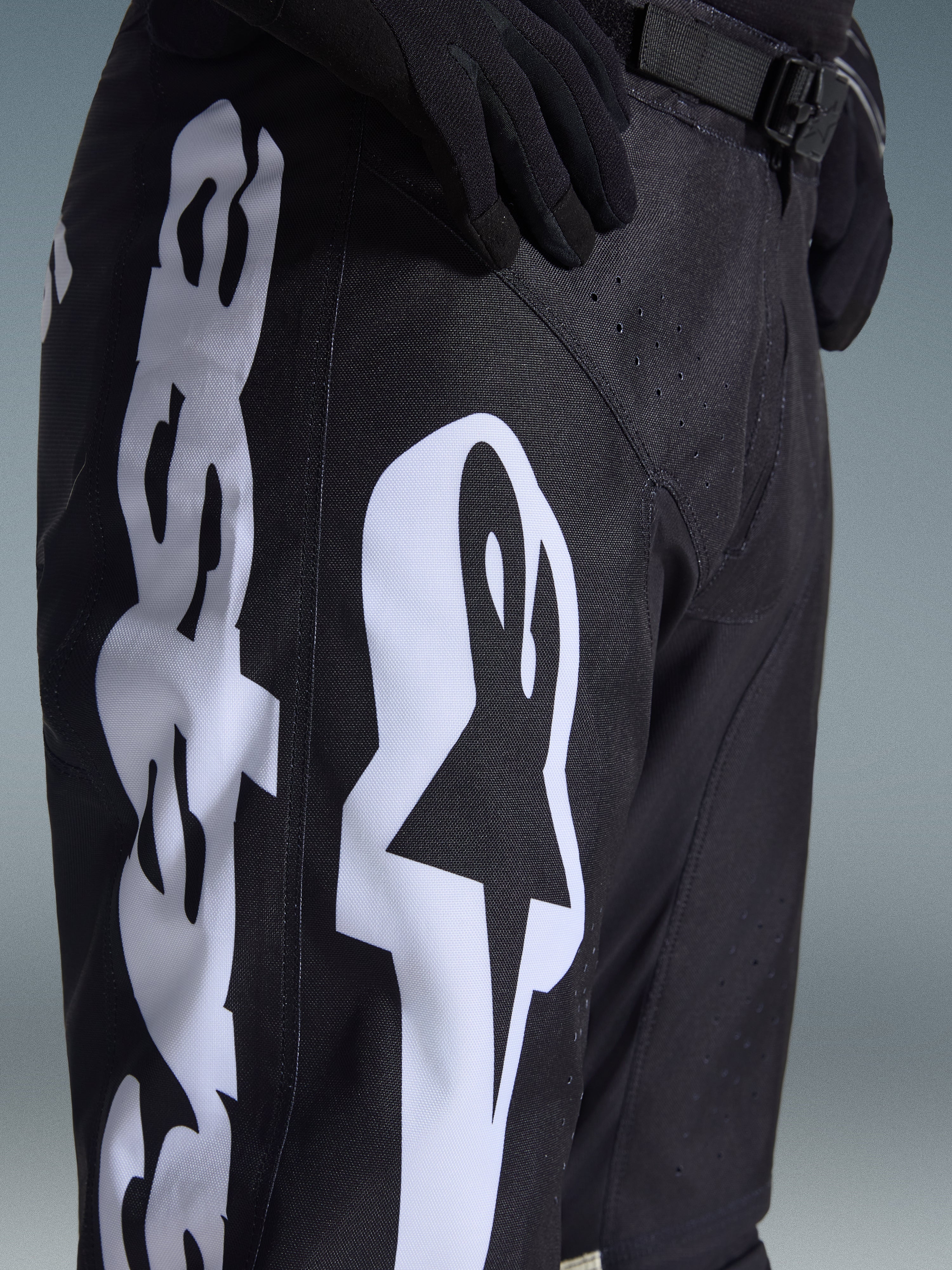 Alpinestars 2026 Racer Air Riway Pants, czarne spodnie motocrossowe z wyraźnym białym logo Alpinestars na udzie, zbliżenie pokazujące techniczny materiał i regulowany pasek w talii, noszone z czarnymi rękawicami do jazdy terenowej