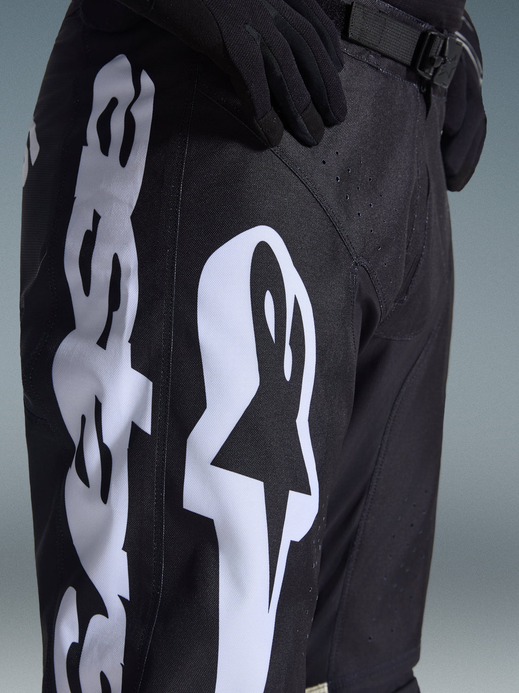 Alpinestars 2026 Racer Air Riway Pants, spodnie motocrossowe, czarno-białe, zbliżenie pokazujące duże białe logo Alpinestars, laserowo wycinane perforacje zapewniające przepływ powietrza oraz regulowaną klamrę w pasie, wytrzymały materiał do jazdy w terenie.