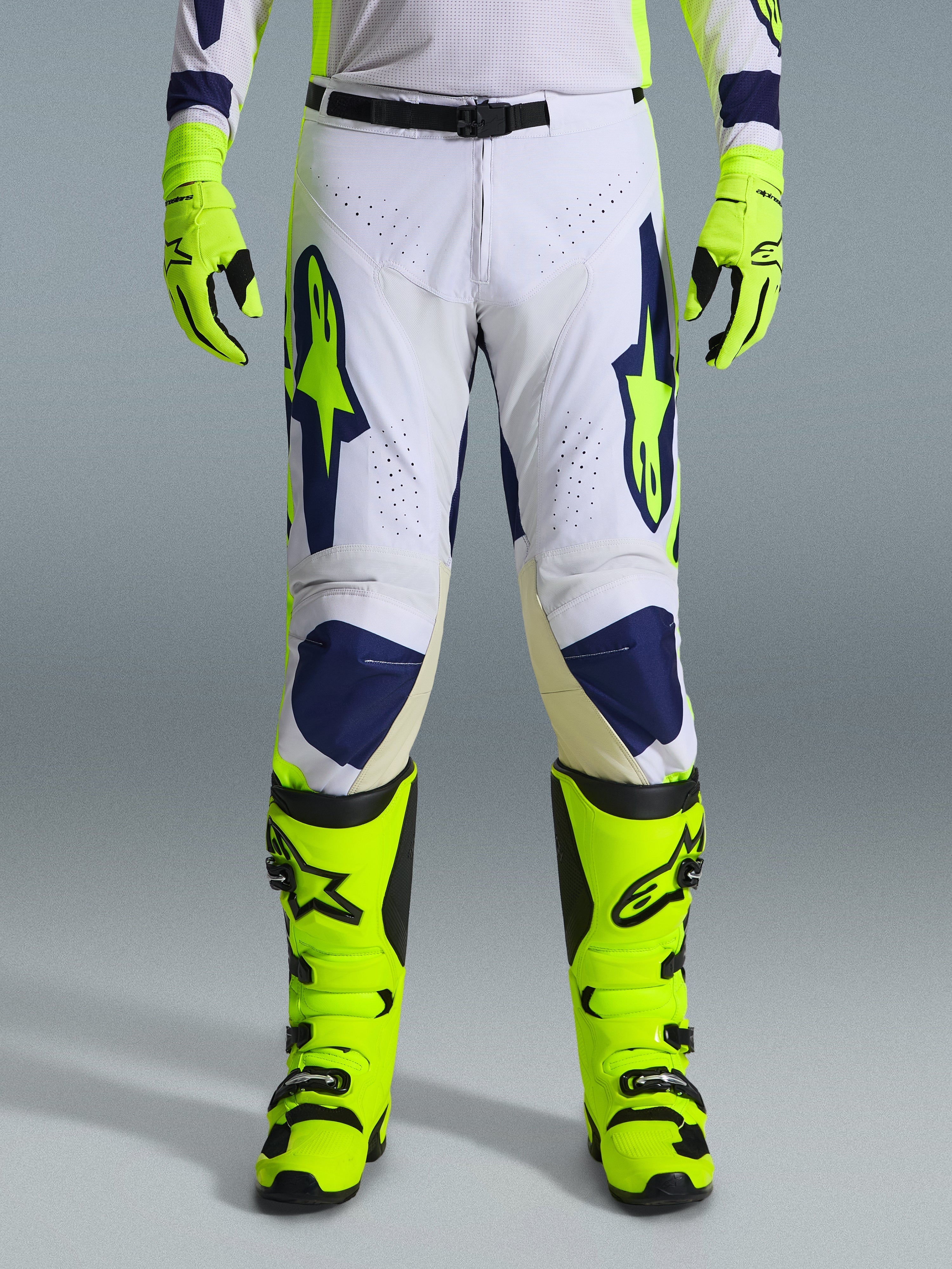 Alpinestars 2026 Racer Air Riway Pants, spodnie motocrossowe, jasnoszare, neonowożółte i niebieskie, noszone z pasującymi neonowożółtymi rękawicami i butami, z perforowanymi przednimi panelami zapewniającymi przepływ powietrza, wyraziste niebieskie i żółte logo, sportowy design do jazdy terenowej