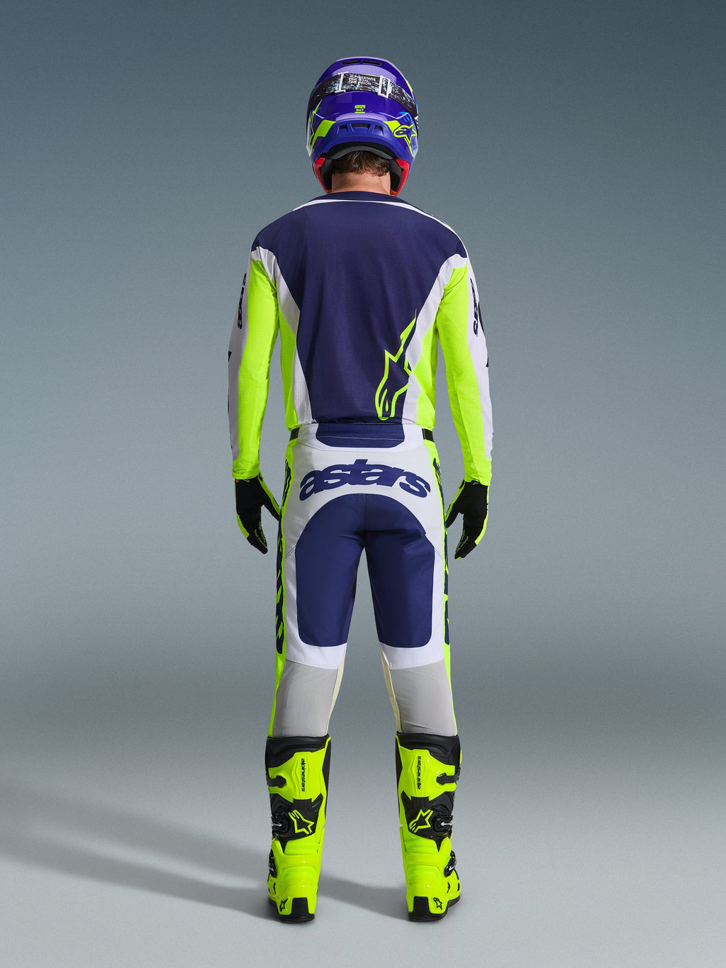 Motocyklista motocrossowy od tyłu w spodniach Alpinestars 2026 Racer Air Riway w kolorze jasnoszarym, żółtym fluo i niebieskim, w zestawie z dopasowaną koszulką i jaskrawożółtymi butami, prezentujący kompletny profesjonalny zestaw odzieży off-road.